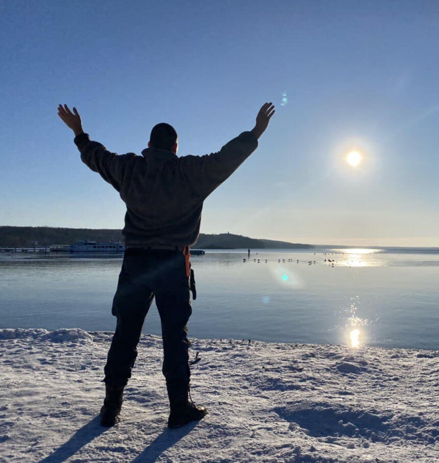 Energiearbeit und Freiheit in der Natur Stralsund „Dennis Jahnke, Coach und energetischer Heiler, steht mit erhobenen Armen und gespreizten Beinen in X-Form am schneebedeckten Seeufer, Sonnenlicht spiegelt sich auf der Wasseroberfläche“